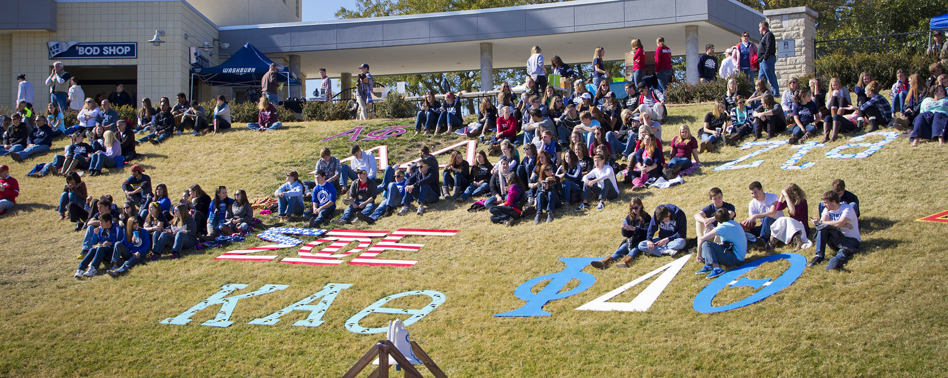 Fraternities and sororities watch a football game from the grassy hill.