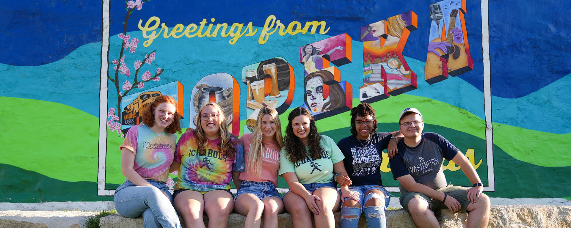 students smiling and wearing colorful tie dye in front of mural in NOTO Topeka, KS
