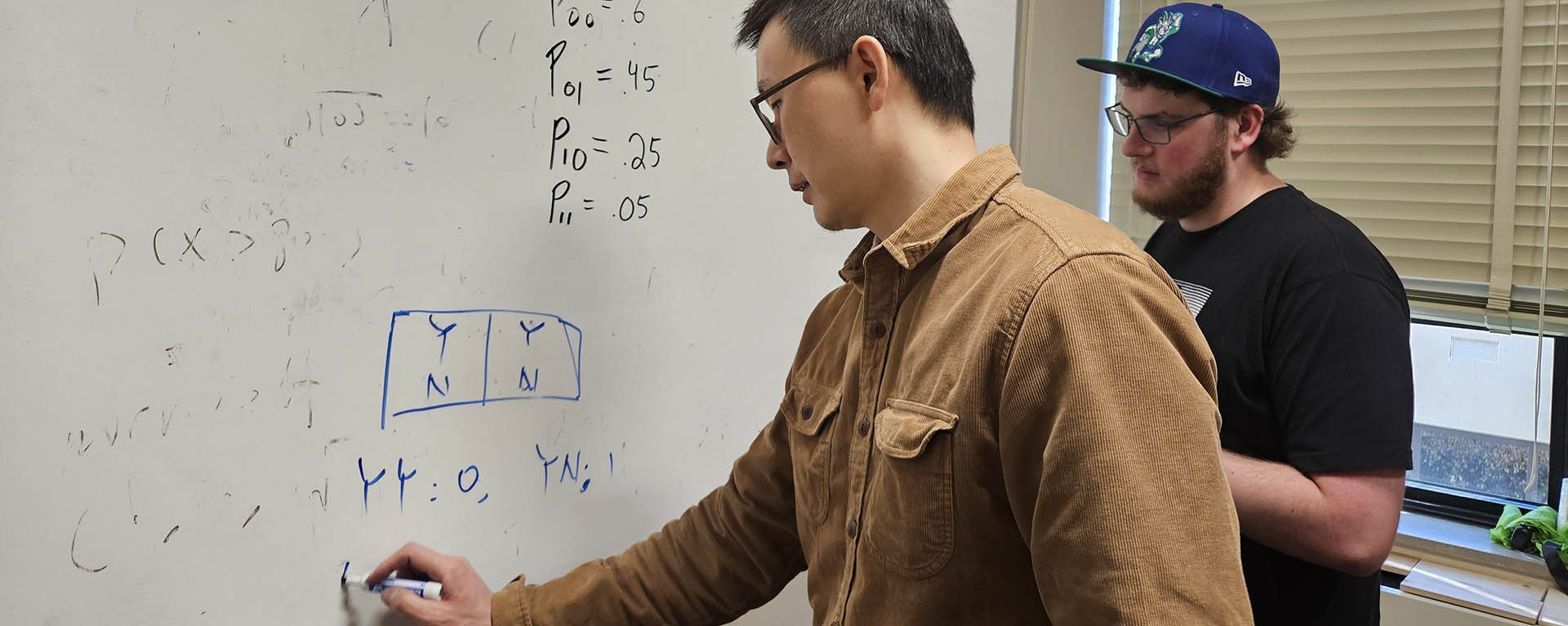 A math professor writes an equation on a white board while a student looks on.