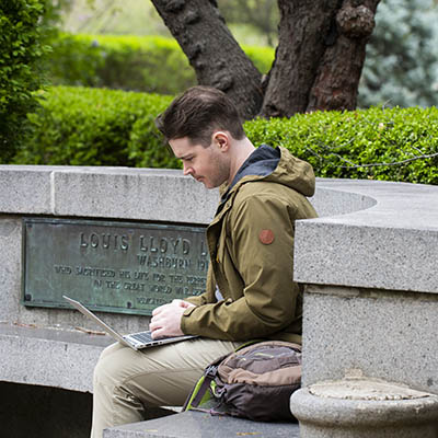 A student works on his laptop while sitting on a concrete bench outside.
