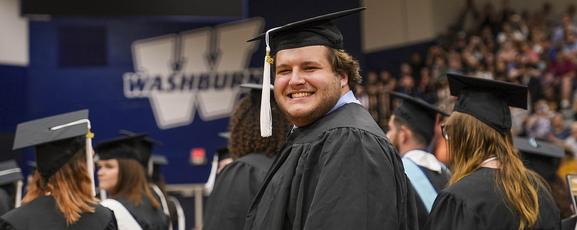 A student smiles widely for a photo in graduation regalia with a Washburn logo in the distance behind him.
