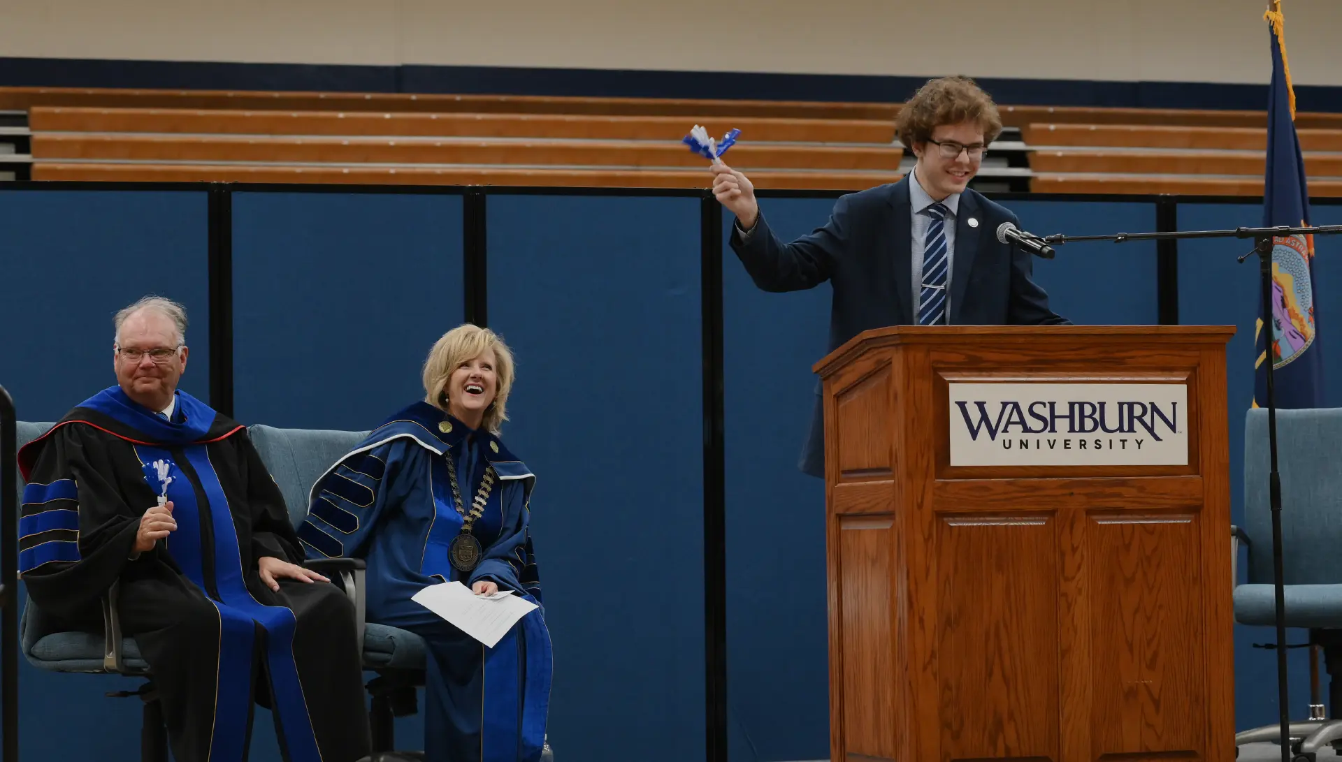 The student body president waves a clapping device while giving a speech at convocation while President Mazachek and Provost Fritch laugh behind.