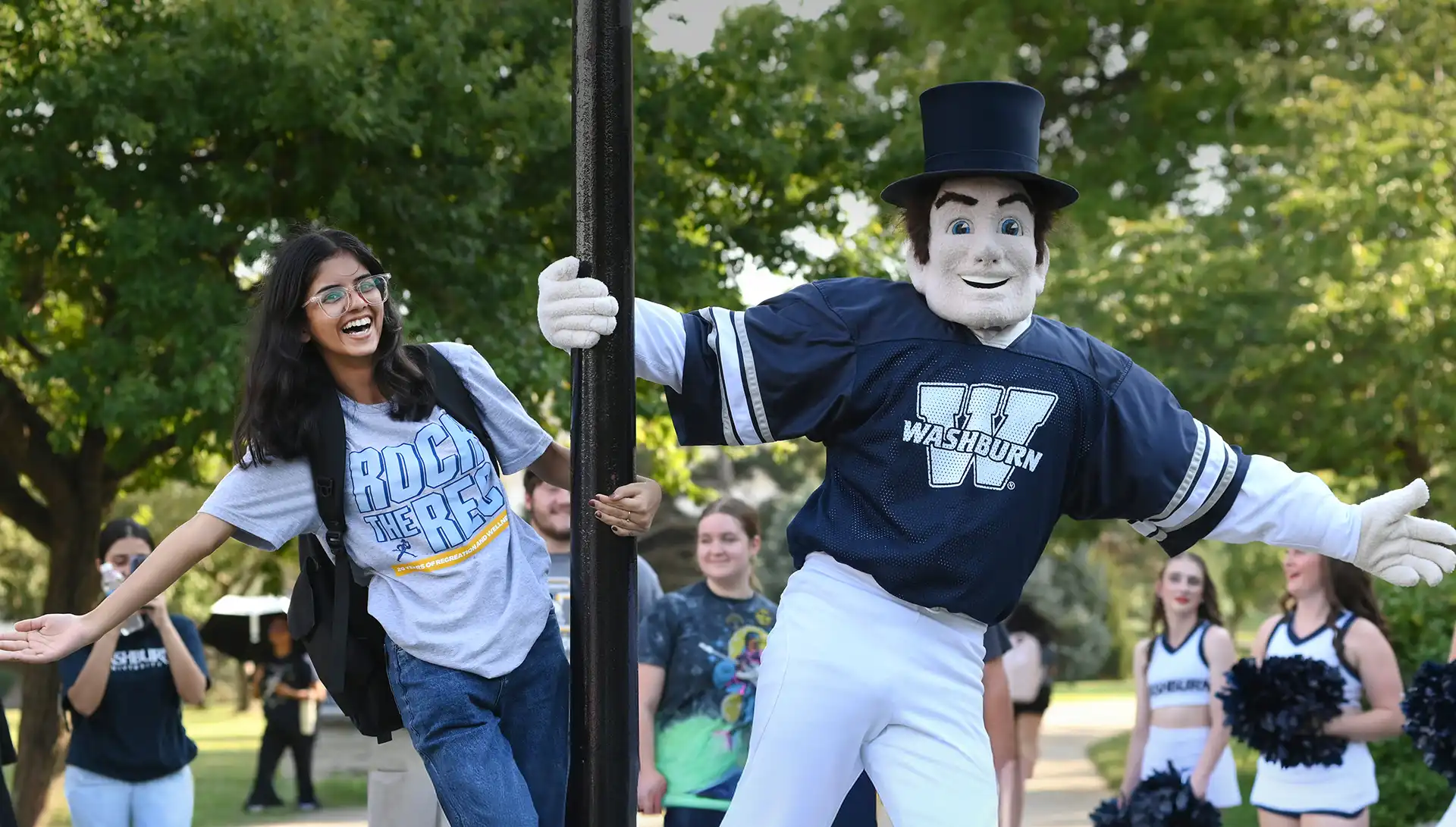 Washburn student and Ichabod mascot swing from lamp post smiling on campus.