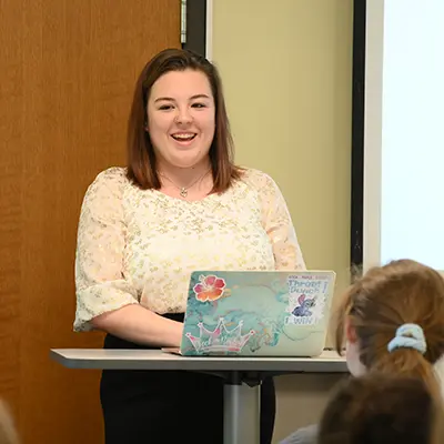 A student smiles while giving a powerpoint presentation at the front of the room.