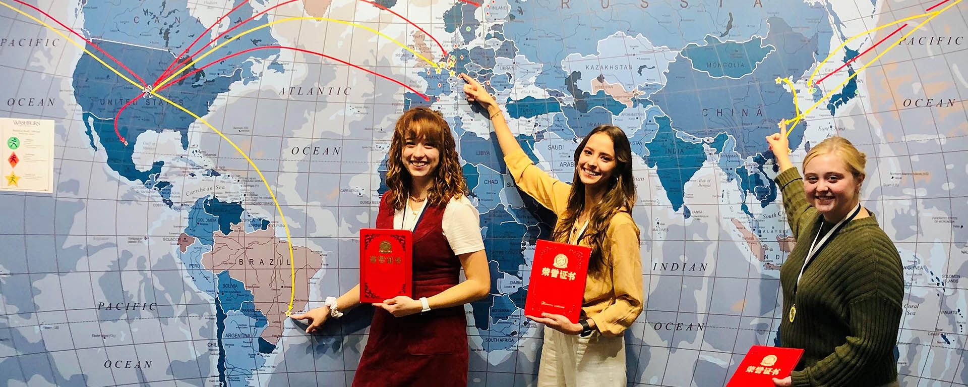 Three business students smile and point to where they studied abroad on a map.