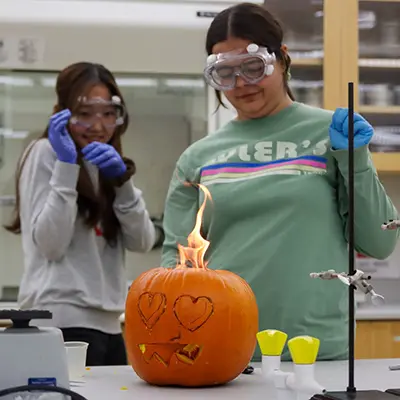 Two chemistry club members perform and experiment in a lab that sets a pumpkin on fire.