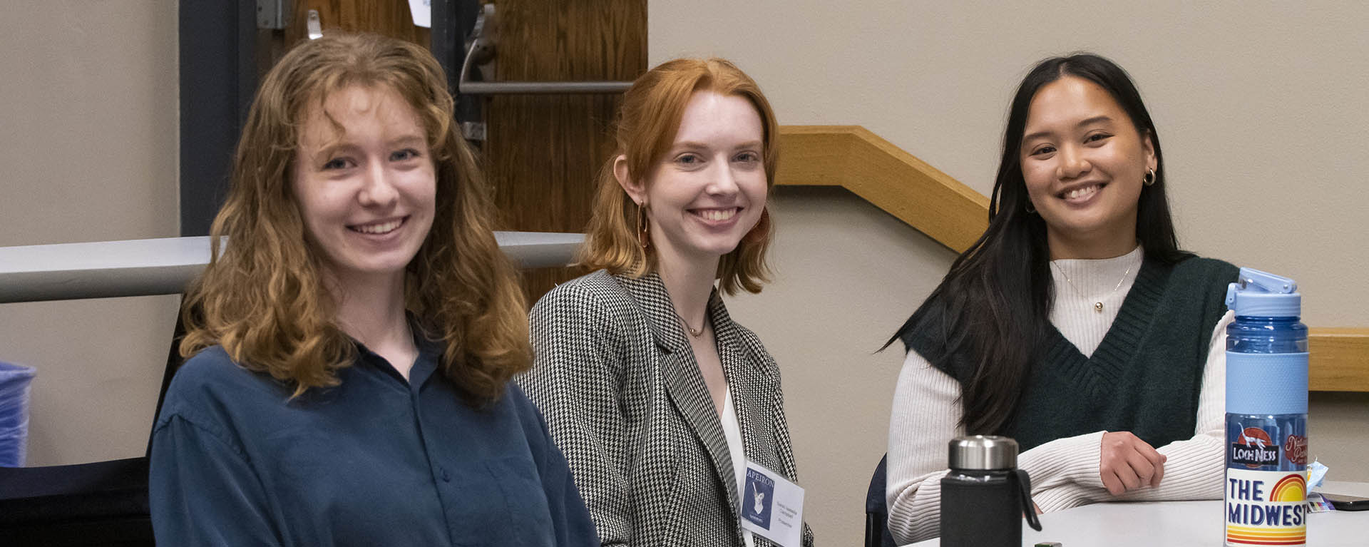 Three students smile while dressed professionally and sitting in a classroom.