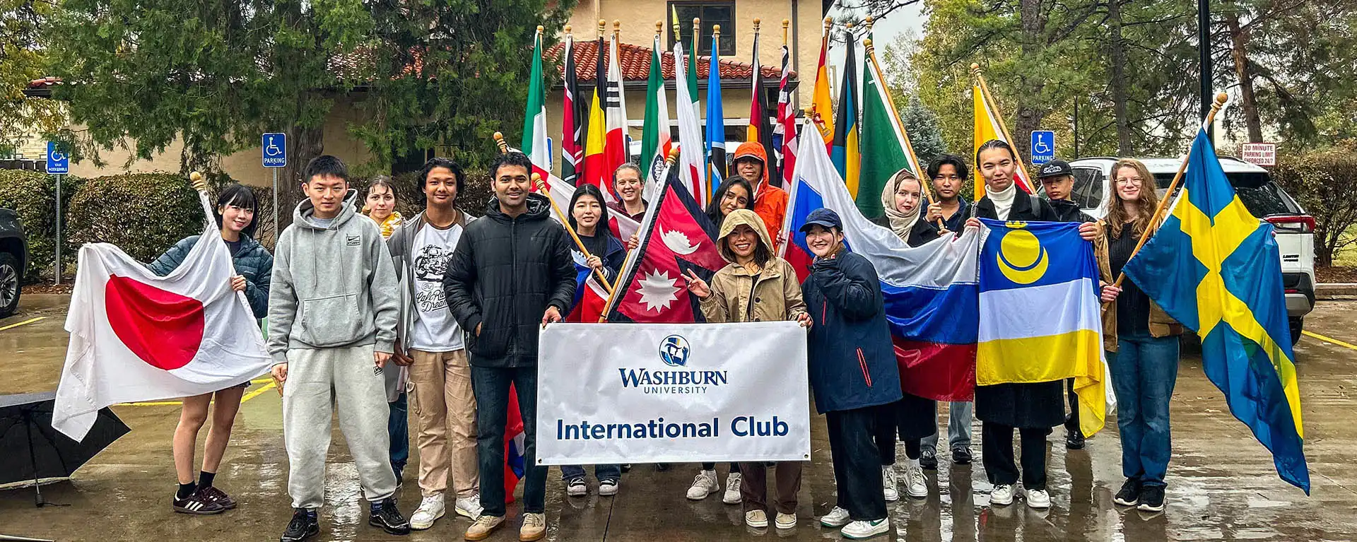 International students smiling and holding flags and 'International Club' sign on campus on rainy day