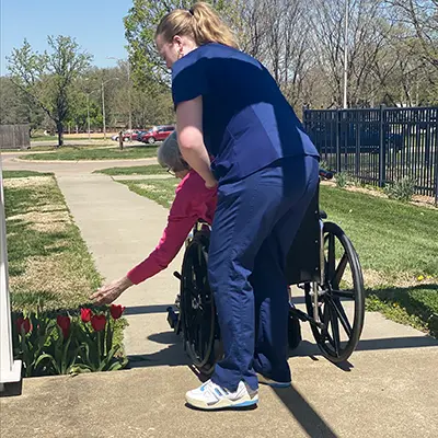 A nursing student pushes a wheelchair while the patient examines flowers along the sidewalk.